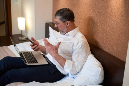 Téléchargez les photos : Homme adulte en tenue de travail assis sur le lit, tenant un téléphone portable et montrant une réaction agressive - en image libre de droit