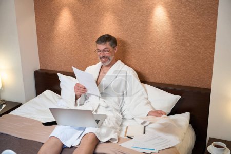 Foto de Varón en albornoz sentado en la cama, sosteniendo la computadora portátil y leyendo documentos en el hotel - Imagen libre de derechos