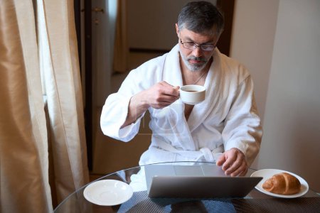 Téléchargez les photos : Homme adulte en peignoir assis sur le canapé, tenant une tasse de thé, buvant et travaillant - en image libre de droit