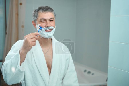 Foto de Varón adulto en albornoz blanco de pie en el baño, mirando en el espejo y afeitado con afeitadora - Imagen libre de derechos