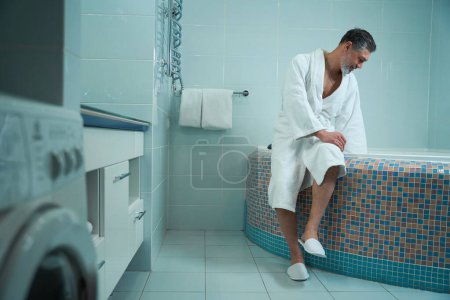 Photo for Adult male in white bathrobe in bathroom sitting on edge of bath and checking water - Royalty Free Image