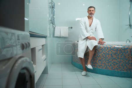 Téléchargez les photos : Homme adulte en peignoir blanc dans la salle de bain assis sur le bord de la baignoire et regardant la caméra - en image libre de droit