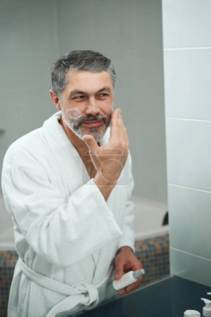 Photo for Adult man in white bathrobe standing in bathroom, looking in mirror and applying cream to face - Royalty Free Image