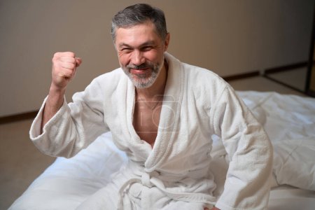 Photo for Adult male in white bathrobe in bedroom sitting on bed and looking at the camera - Royalty Free Image