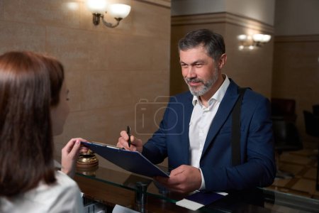 Téléchargez les photos : Homme en vêtements de travail debout près de la réception et regardant administrateur, écrit sur le document - en image libre de droit