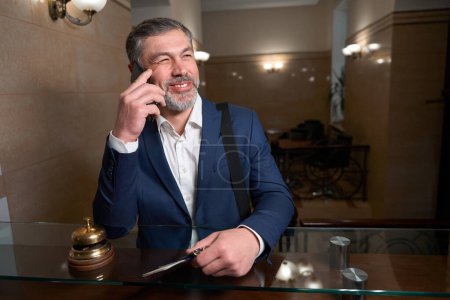 Téléchargez les photos : Homme en tenue de travail debout près de la réception, tenant passeport et clés et parlant au téléphone - en image libre de droit