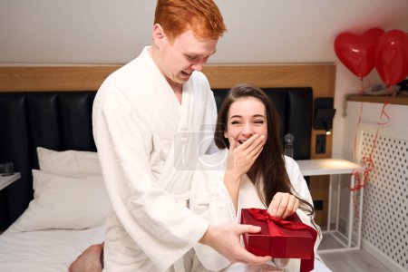 Téléchargez les photos : Jeune homme félicite sa mignonne petite amie le jour de la Saint-Valentin, un cadeau dans une boîte rouge - en image libre de droit