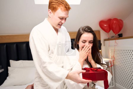 Téléchargez les photos : Un bel homme roux souhaite à sa charmante femme une joyeuse Saint Valentin, un cadeau dans une boîte rouge - en image libre de droit