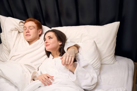 Foto de El pelirrojo y su novia con albornoces yacen en una cama grande, una pareja descansa en un hotel - Imagen libre de derechos
