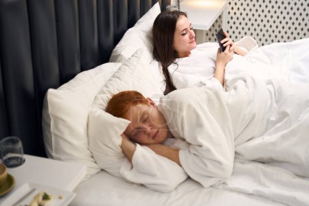 Téléchargez les photos : Belle jeune femme est couchée au lit avec un téléphone, son mari dort doucement à proximité - en image libre de droit