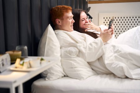 Photo for Cheerful couple is resting in a hotel room and chatting on phones, they are in a large and soft bed - Royalty Free Image