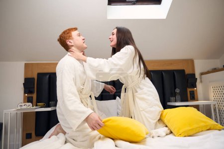 Foto de Los cónyuges jóvenes comenzaron una pelea de almohadas cómicas en la cama, los chicos alquilan una habitación de hotel - Imagen libre de derechos