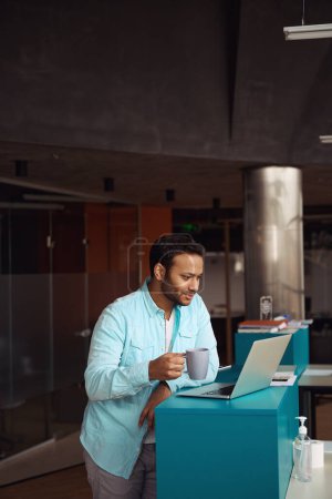 Téléchargez les photos : Jeune homme debout près de l'ordinateur et tenant la tasse dans sa main dans le bureau - en image libre de droit