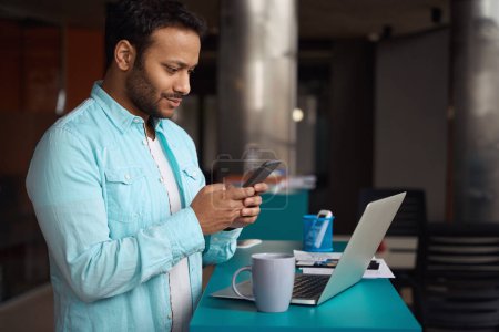Téléchargez les photos : Homme debout à table avec ordinateur portable et tapant sur le téléphone dans le centre d'affaires - en image libre de droit