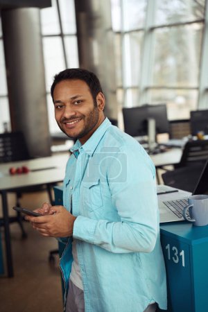 Foto de Hombre sonriente parado en el escritorio con taza y computadora y escribiendo en el teléfono inteligente en la oficina - Imagen libre de derechos
