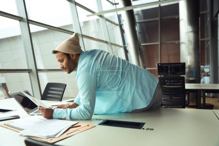 Téléchargez les photos : Jeune homme en chapeau assis sur le bureau derrière l'ordinateur et le plan de travail de lecture dans le centre d'affaires - en image libre de droit
