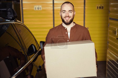 Téléchargez les photos : Homme souriant debout près de la voiture dans la salle de stockage avec la boîte dans ses mains - en image libre de droit