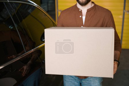 Téléchargez les photos : Jeune homme barbu tenant une boîte dans ses mains près d'une machine dans la salle de stockage - en image libre de droit
