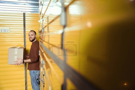 Téléchargez les photos : Agréable adulte mâle gardant la boîte dans ses mains tout en se tenant dans l'entrepôt - en image libre de droit