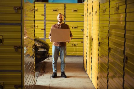 Photo for Pleased groown male standing in the warehouse while carrying the cardboard box - Royalty Free Image