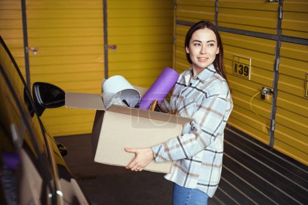 Téléchargez les photos : Femme adulte souriante debout dans un entrepôt près de la voiture tout en gardant une boîte avec des choses - en image libre de droit