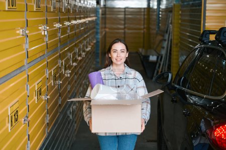 Photo for Happy young female standing in the storehouse near her car while keeping a box with stuff - Royalty Free Image