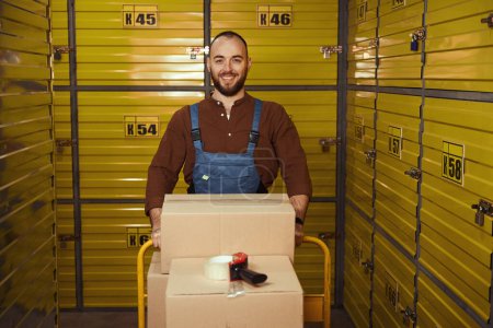 Téléchargez les photos : Un homme heureux tenant la chargeuse pleine de boîtes en carton debout dans la chambre forte - en image libre de droit