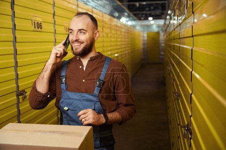 Téléchargez les photos : Homme souriant debout près des boîtes dans la salle de stockage tout en parlant au téléphone - en image libre de droit