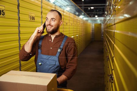 Téléchargez les photos : Homme joyeux debout derrière les casiers de stockage tout en ayant une conversation sur smartphone dans un entrepôt - en image libre de droit