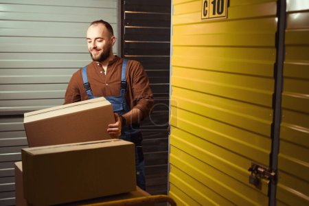 Téléchargez les photos : Jeune homme souriant tenant une boîte en carton debout dans un entrepôt - en image libre de droit