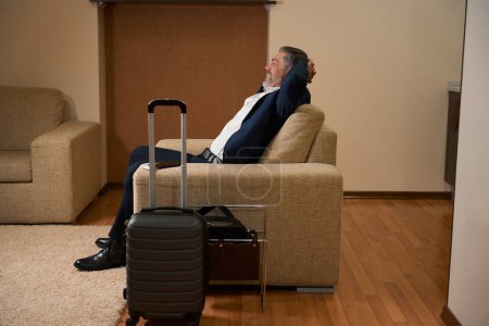Foto de Hombre feliz en traje de negocios junto a la maleta está descansando en el hotel - Imagen libre de derechos