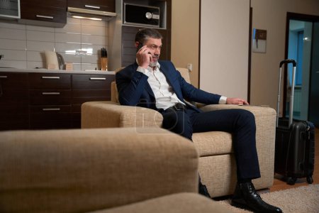 Foto de Hombre adulto en traje de negocios sentado en sillón y hablando por teléfono en la habitación del hotel - Imagen libre de derechos