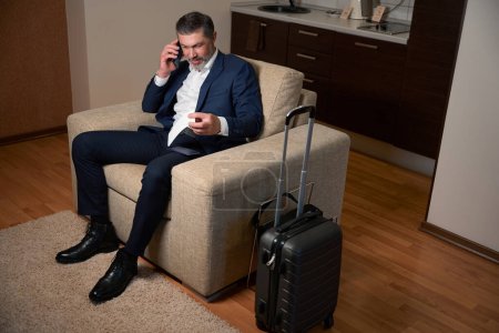 Photo for Man siting in armchair and calling on phone from business card in the hotel - Royalty Free Image