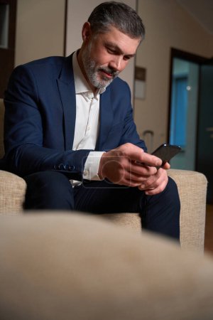 Foto de Hombre de traje de negocios sentado en silla y escribiendo por teléfono en el hotel - Imagen libre de derechos