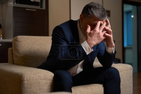 Foto de Hombre adulto en ropa de oficina sentado en silla y cubrir su cara con las manos en el motel - Imagen libre de derechos