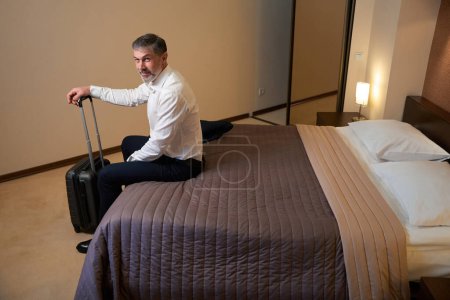 Photo for Adult male resting on the bed and holding the handle of the suitcase in the hostel - Royalty Free Image