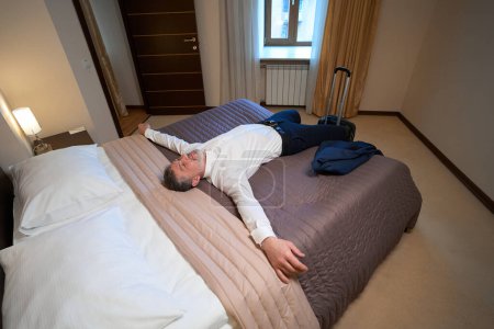 Photo for Adult male in office clothes resting on bed near suitcase in the hotel - Royalty Free Image