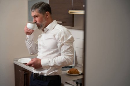 Foto de Hombre en ropa de oficina de pie cerca de la mesa con la taza en la mano en el hotel - Imagen libre de derechos