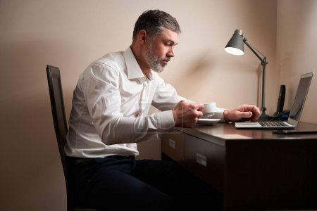 Foto de Hombre adulto sentado en la silla en la mesa delante de la computadora y mirando en la taza en el hotel - Imagen libre de derechos