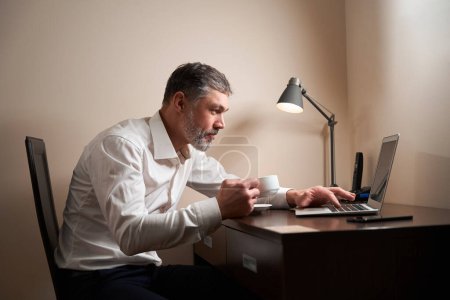 Téléchargez les photos : Homme assis sur une chaise à table avec une tasse dans la main et regardant dans un ordinateur portable - en image libre de droit