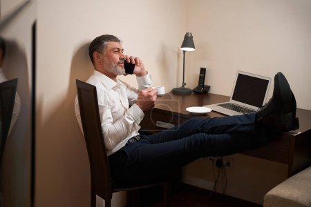 Téléchargez les photos : Homme adulte assis sur une chaise devant un ordinateur portable, buvant du thé et utilisant un smartphone dans l'hôtel - en image libre de droit