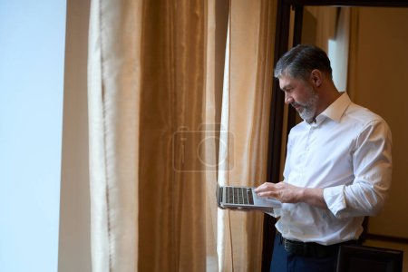 Photo for Adult male in business suit standing near window and using computer in the hostel - Royalty Free Image