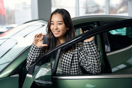 Foto de Joven cliente femenino se ve fuera de coche nuevo y tiene una llave en las manos, una mujer con ropa elegante - Imagen libre de derechos