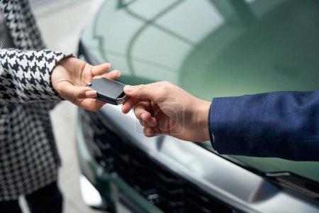 Frau nimmt Mann im Businessanzug Autoschlüssel ab, Menschen stehen vor glänzendem Neuwagen