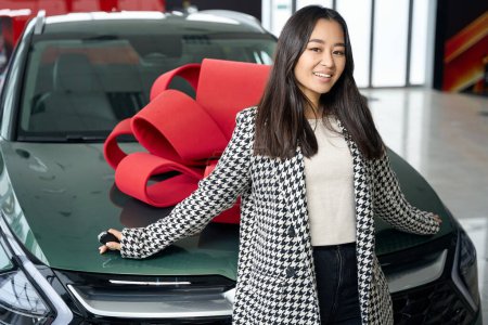 Téléchargez les photos : Femme asiatique souriante se tient dans un concessionnaire automobile sur le fond d'une voiture avec un arc cadeau rouge, femelle en vêtements décontractés - en image libre de droit