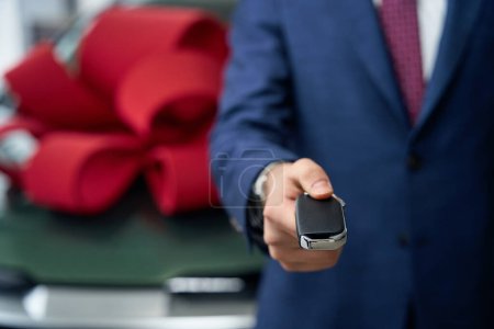 Téléchargez les photos : Homme en costume d'affaires tend la main avec la clé de voiture, en arrière-plan est voiture avec un arc cadeau rouge - en image libre de droit