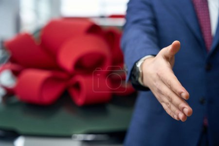 Foto de Hombre en un traje de negocios extiende su mano para saludar, en el fondo un coche con un arco de regalo rojo - Imagen libre de derechos