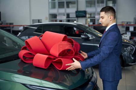 Foto de Bonito hombre sonriente adjunta un arco de regalo rojo a la capucha del coche, macho en un concesionario de automóviles - Imagen libre de derechos