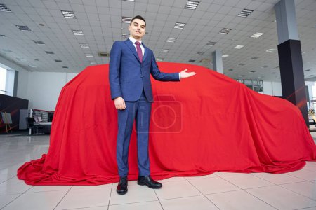 Foto de Hombre en un traje de negocios se pone delante de un coche cubierto con un paño rojo, el gerente apunta al coche con la mano - Imagen libre de derechos