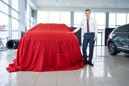 Photo for Car dealership consultant stands against the background of a car covered with a red cloth, viewports in a car dealership - Royalty Free Image
